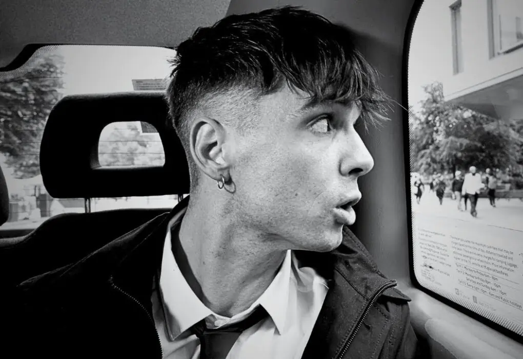 Retrato en blanco y negro de Ren Gill dentro de un taxi, mirando pensativo por la ventana mientras lleva una camisa blanca y corbata aflojada.