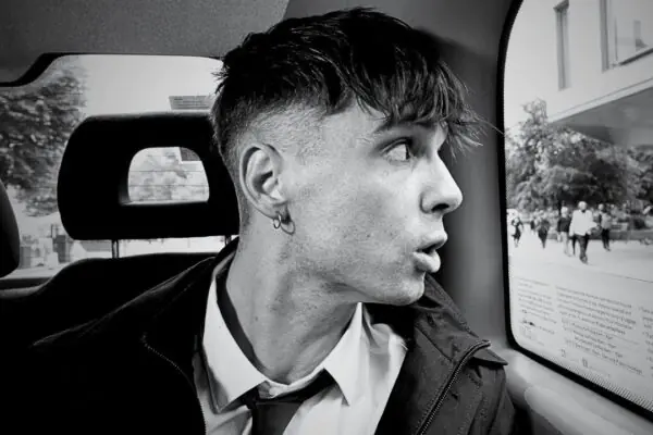 Retrato en blanco y negro de Ren Gill dentro de un taxi, mirando pensativo por la ventana mientras lleva una camisa blanca y corbata aflojada.