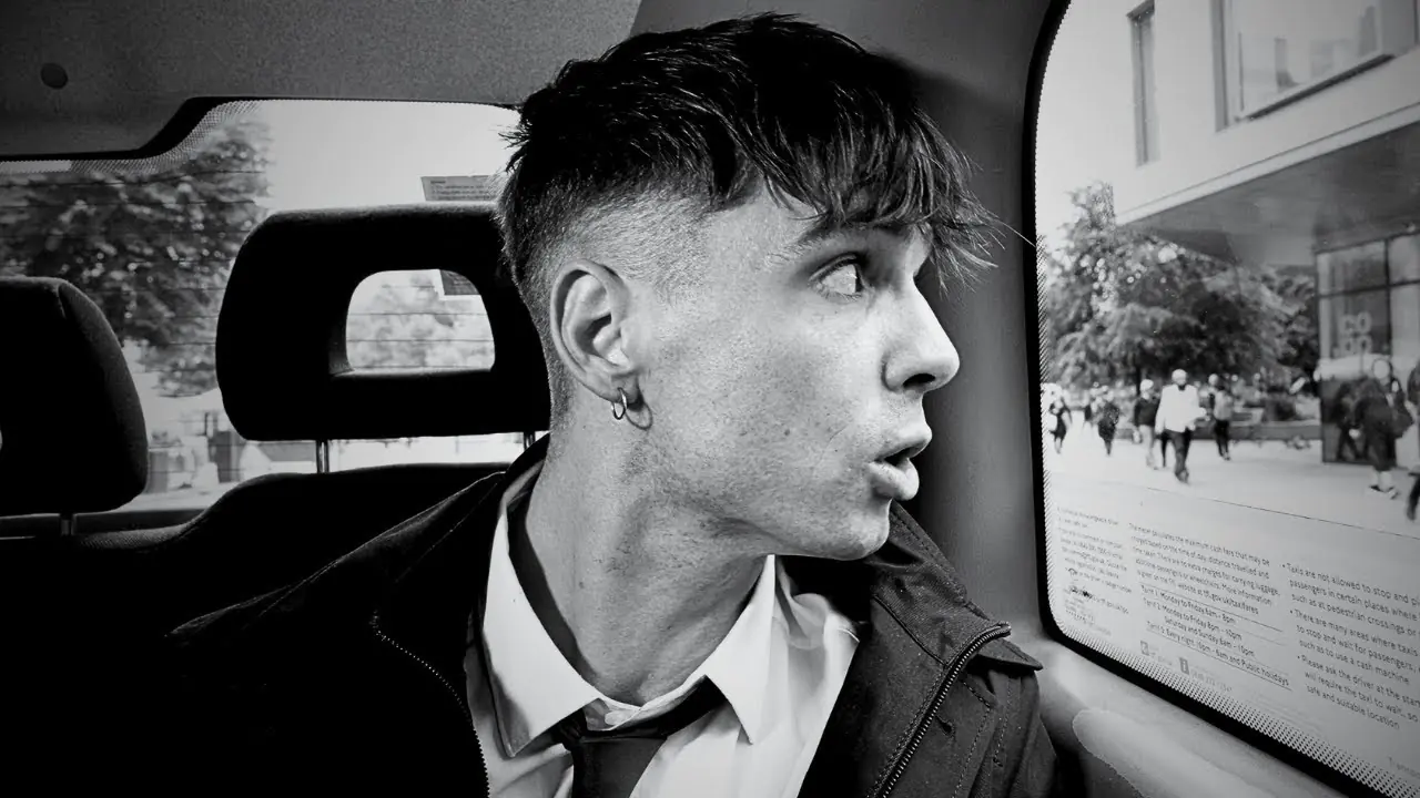 Retrato en blanco y negro de Ren Gill dentro de un taxi, mirando pensativo por la ventana mientras lleva una camisa blanca y corbata aflojada.